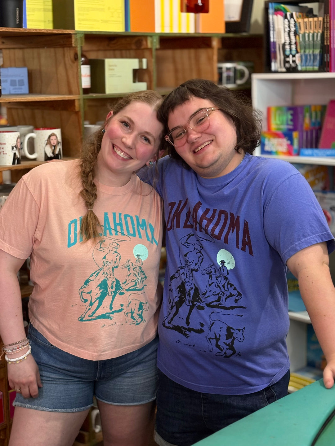 Two people smiling and posing together in a shop, wearing matching graphic t-shirts with 'OKLAHOMA' printed on them. The person on the left wears a peach-pink t-shirt with teal lettering and illustration, paired with denim shorts.