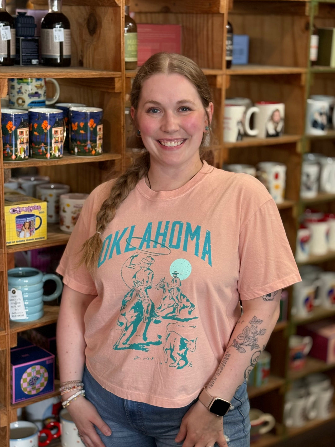 A smiling woman with a braid stands in a shop in front of wooden shelves filled with mugs and bottles, wearing a peach-colored graphic t-shirt with 'OKLAHOMA' printed in teal.