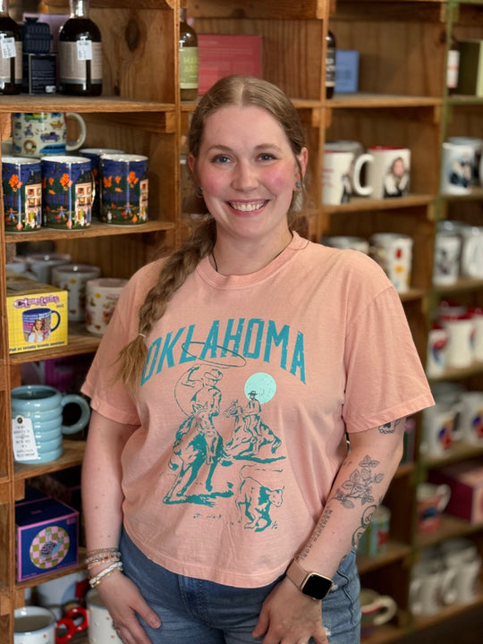 A smiling woman with a braid stands in a shop in front of wooden shelves filled with mugs and bottles, wearing a peach-colored graphic t-shirt with 'OKLAHOMA' printed in teal.