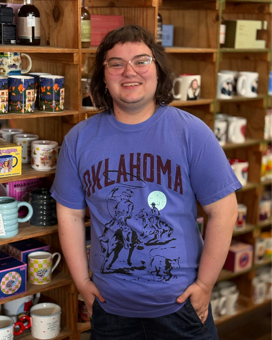 A person standing in a shop in front of wooden shelves filled with mugs and bottles, wearing a blue graphic t-shirt that says 'OKLAHOMA' with a cowboy and horse design.