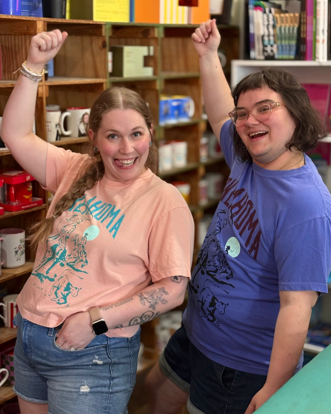 Two people standing in a shop or cafe setting, both wearing graphic t-shirts with 'Oklahoma' printed on them. The person on the left wears a peach-pink t-shirt and denim shorts, while the person on the right wears a blue t-shirt and dark shorts.