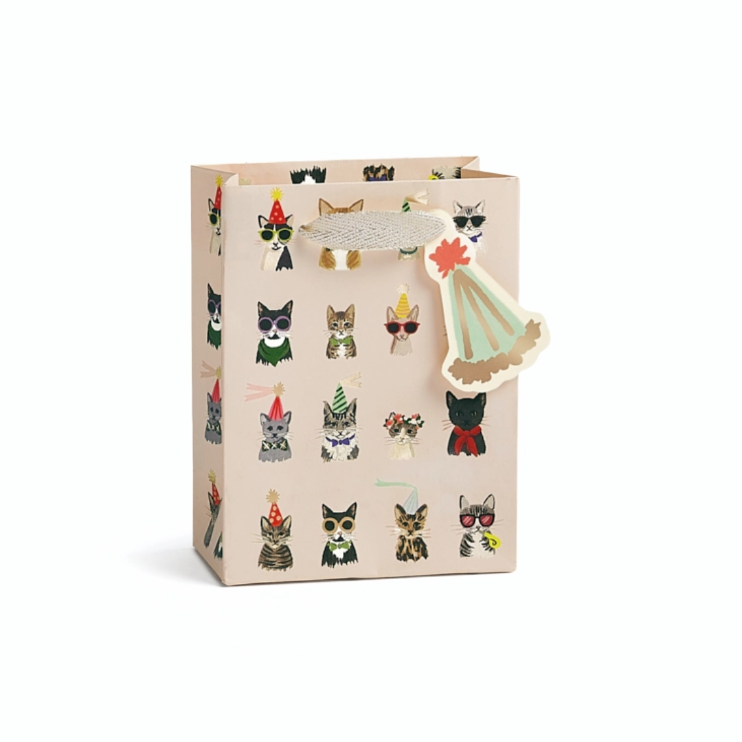 A beige gift bag decorated with a pattern of illustrated cats in various party hats, featuring a silver metallic handle and a decorative paper tag.