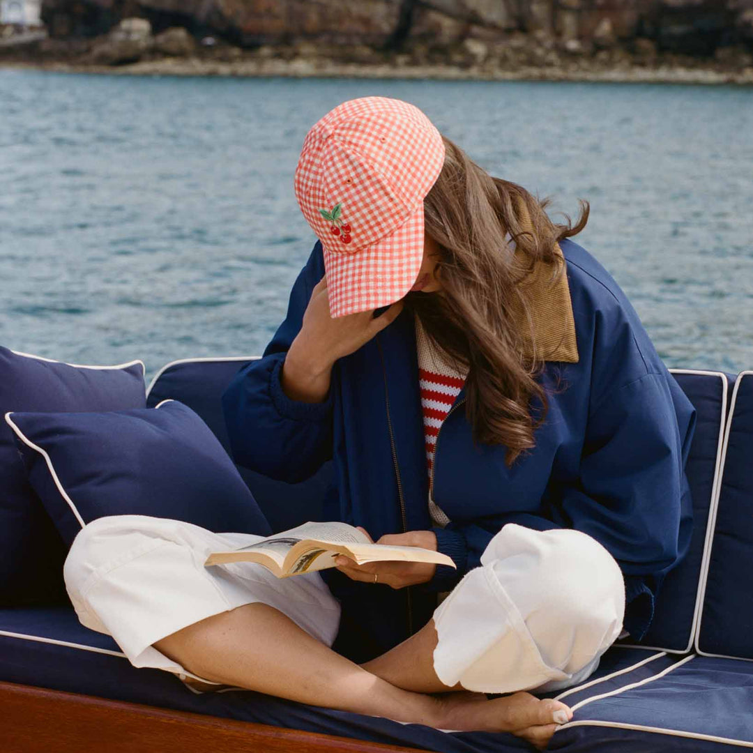 Person reading a book on a boat with a scenic background while wearing a red gingham hat with a cherry design.