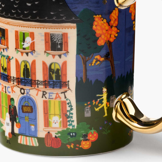 A close-up of a ceramic mug featuring a colorful Halloween illustration with a haunted house, pumpkins, trees, and a gold handle.