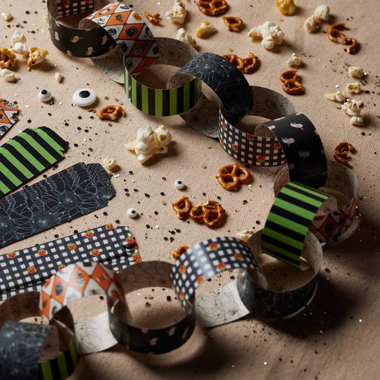 A Halloween-themed paper chain made from patterned paper rolls, styled on a brown kraft paper background with scattered pretzels, popcorn, and googly eyes.