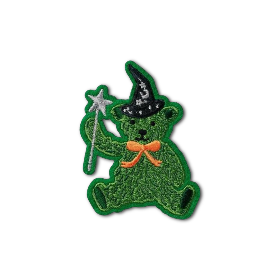 An embroidered patch of a green teddy bear dressed as a wizard, wearing a black pointed hat with white stars and holding a silver wand, set against a plain white background.