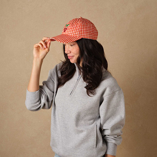 Person wearing a gray hoodie and red checkered cap with an embroidered cherries design against a beige background