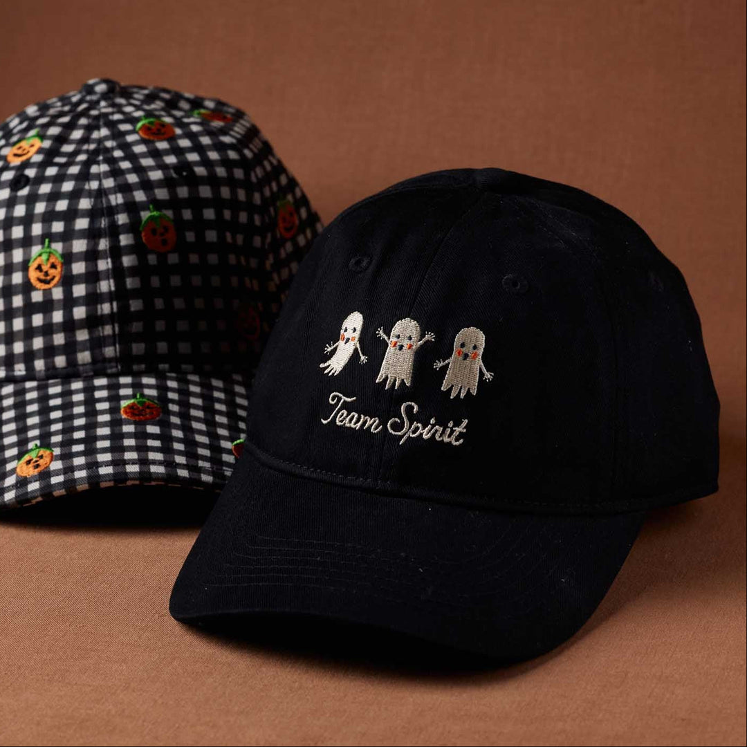 Two baseball caps on a warm brown studio background. The front cap is black with embroidered white ghosts and the words 'Team Spirit.