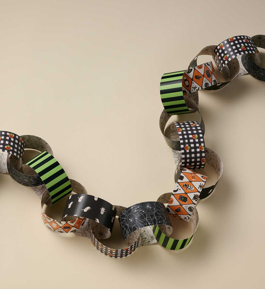 A handmade paper chain garland made of patterned paper rolls, featuring Halloween designs like stripes, checkers, spiderwebs, and pumpkins on a beige background.
