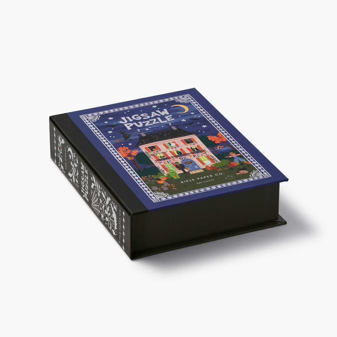 A boxed jigsaw puzzle on a clean white background. The box is black with ornate white side patterns, and the top lid shows a colorful illustration of a house at night under a crescent moon, with the words 'Jigsaw Puzzle' visible.