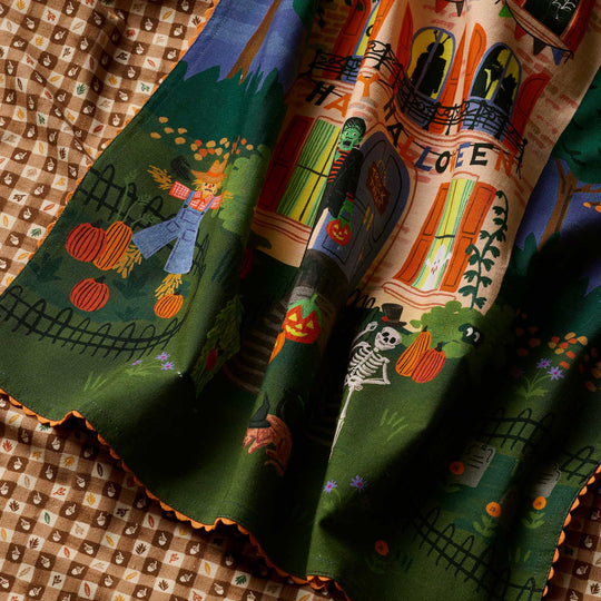 A close-up of a tea towel featuring a Halloween design with a haunted house, pumpkins, a skeleton, and a scarecrow on a green background, draped over a brown checkered fabric.