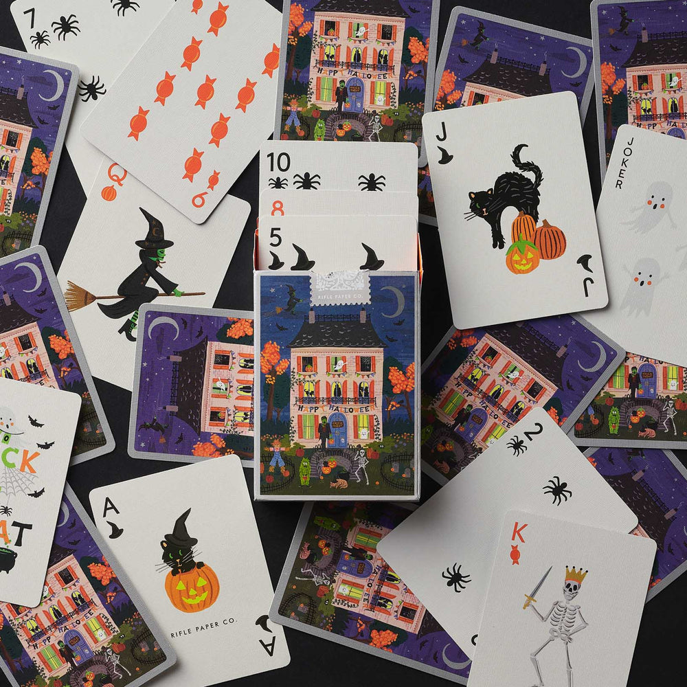 A flat-lay arrangement of Halloween-themed playing cards on a dark background, featuring illustrated houses, bats, witches, pumpkins, ghosts, and jack-o'-lanterns.