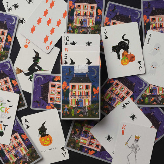 A flat-lay arrangement of Halloween-themed playing cards on a dark background, featuring illustrated houses, bats, witches, pumpkins, ghosts, and jack-o'-lanterns.