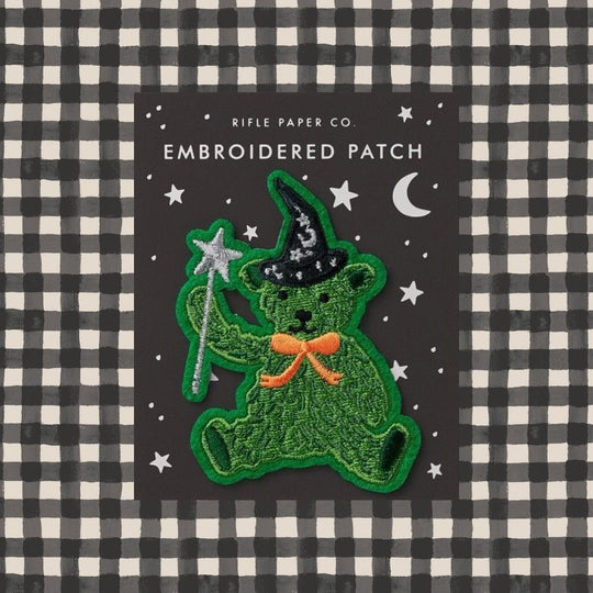 An embroidered patch featuring a green teddy bear dressed as a witch with a black hat, silver wand, and orange bow, displayed on a dark backing card with white text reading 'RIFLE PAPER CO. EMBROIDERED PATCH' against a black and white gingham background.