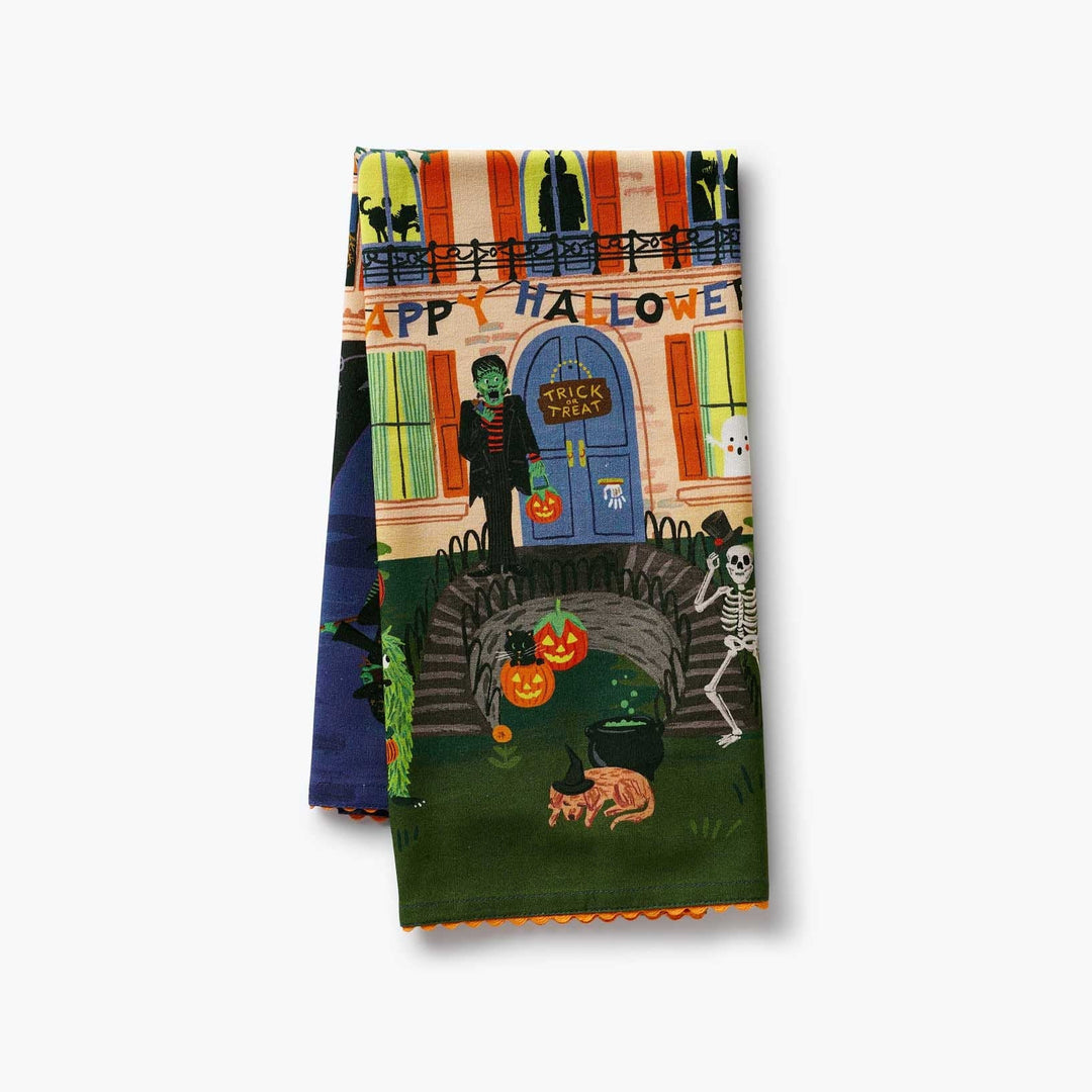 A folded Halloween-themed kitchen towel featuring a colorful illustration of a haunted house, pumpkins, a skeleton, a cat, and a green character in front of a blue door with a 'Trick or Treat' sign.