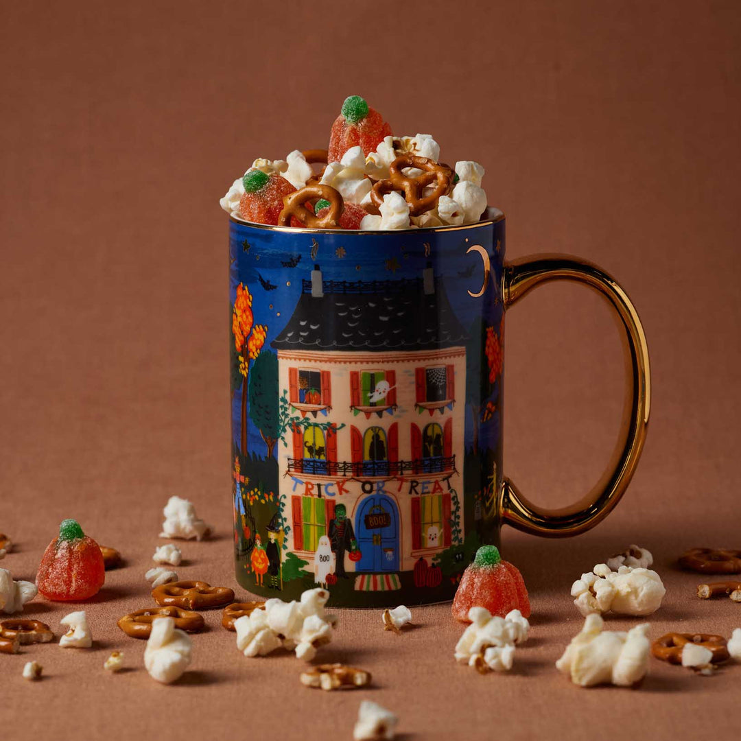A Halloween-themed ceramic mug with a gold handle, filled with popcorn, pretzels, and gummy candies, set against a solid brown background with scattered snacks around it.