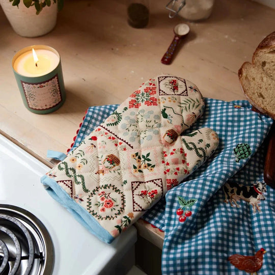A rustic patterned oven mitt sits on a wooden surface with a lit candle.