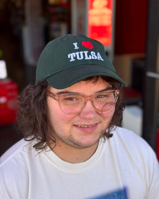 A person smiles while wearing a green hat with the text "I ❤️ TULSA".