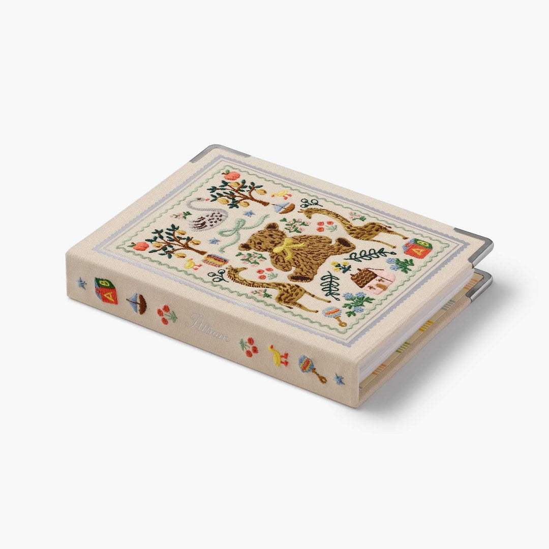 A decorative hardcover photo album book with a cream-colored cover featuring a colorful embroidery of a teddy bear, apples, birds, and small motifs. The album is shown at an angle on a clean, light gray background.