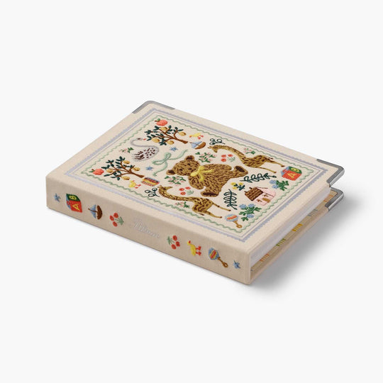 A decorative hardcover photo album book with a cream-colored cover featuring a colorful embroidery of a teddy bear, apples, birds, and small motifs. The album is shown at an angle on a clean, light gray background.