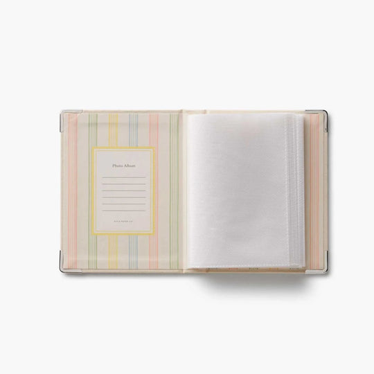 An open photo album with a pastel striped cover in pink, yellow, green, and blue, featuring a photo frame insert on the left and white photo sleeves on the right, set against a plain white background.