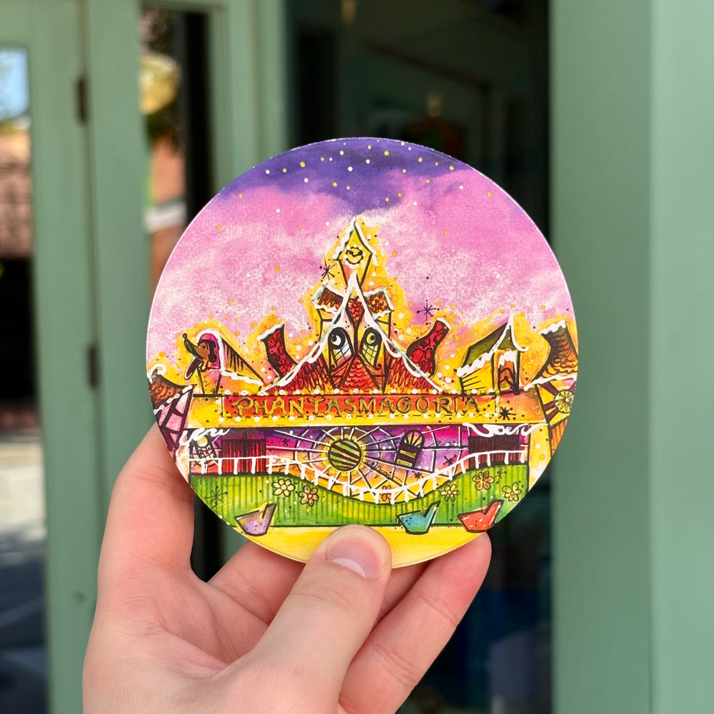 A hand holds a round, colorful printed coaster featuring a whimsical castle illustration with the text 'PHANTASMAGORIA' across the middle, set against a blurred outdoor background.