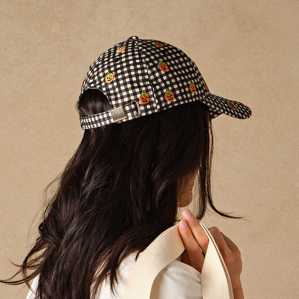 A woman shown from the back wearing a black and white gingham baseball cap with small orange pumpkin motifs, holding a cream canvas tote bag against a textured beige background.
