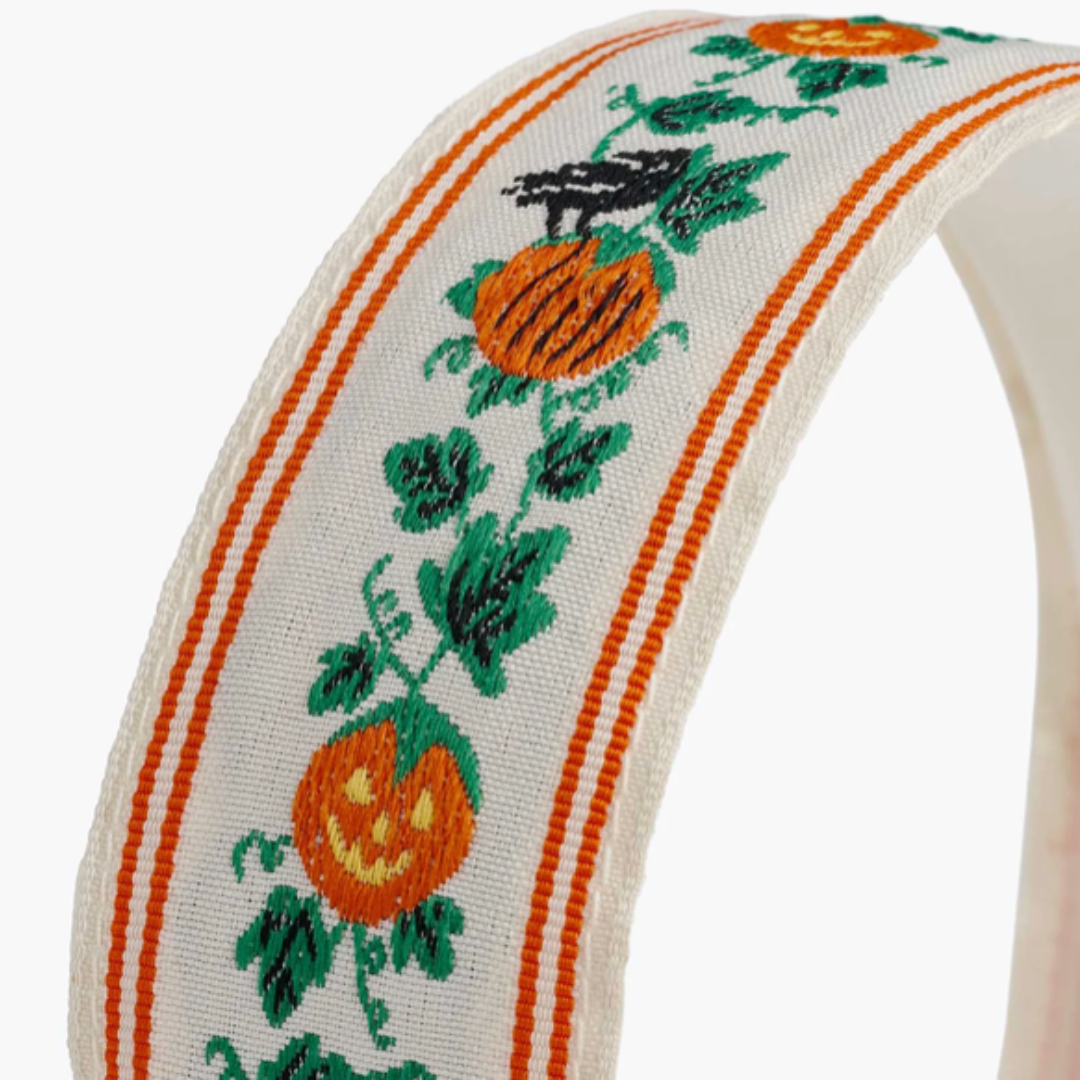 A close-up of a white headband featuring an embroidered orange and yellow pumpkin vine with green leaves, bordered by orange stripes.