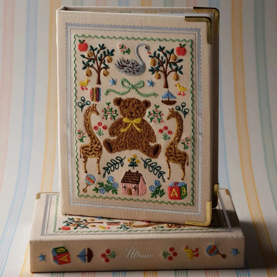 An embroidered baby album with a teddy bear design. The cover features a colorful embroidered scene with a teddy bear, giraffes, trees, and a swan on a cream background.
