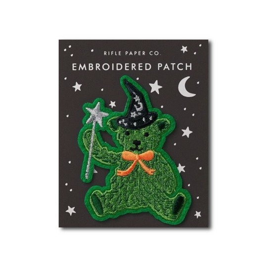 An embroidered patch of a green teddy bear wearing a black witch hat and holding a silver wand, displayed on its dark gray backing card with white stars and the text 'RIFLE PAPER CO. EMBROIDERED PATCH'.