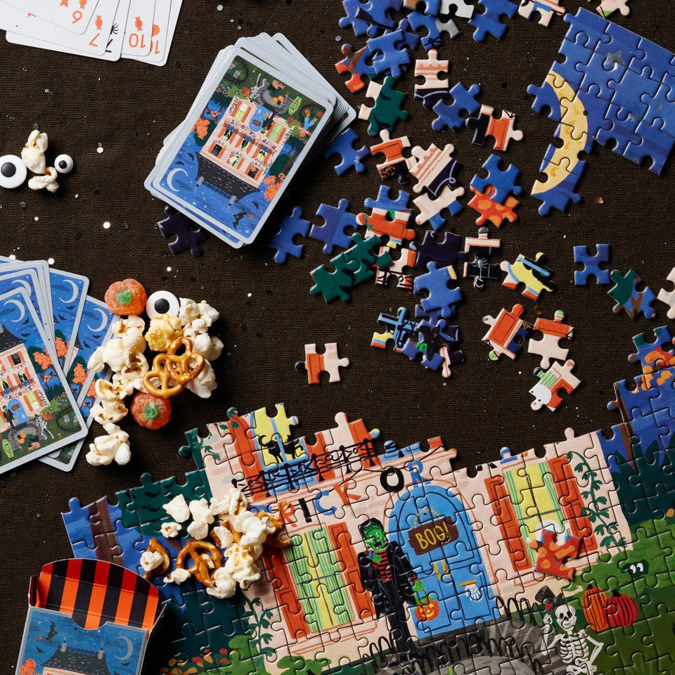 A styled flat-lay of jigsaw puzzle pieces, completed and partially assembled puzzles, playing cards, and popcorn scattered on a dark tabletop.