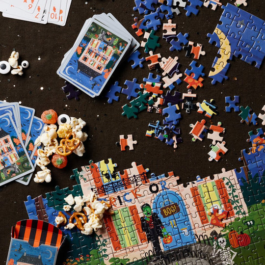 A styled flat-lay of jigsaw puzzle pieces, completed and partially assembled puzzles, playing cards, and popcorn scattered on a dark tabletop.