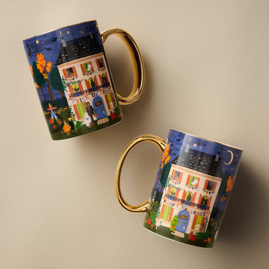 Two ceramic mugs with gold handles, featuring a whimsical illustration of a haunted house at night under a starry sky, displayed on a neutral beige background.