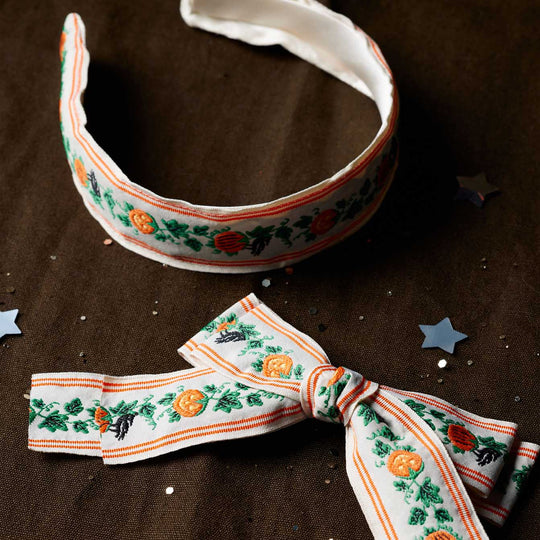 A fabric headband and matching bow featuring a repeating pattern of orange pumpkins, green holly leaves, and black bats on a white background, styled on a dark brown textured surface with scattered silver star confetti.