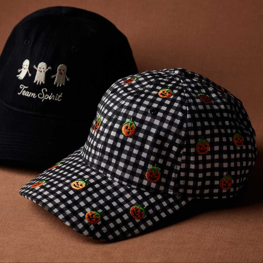 Two baseball caps on a warm brown studio background. The foreground cap is black and white gingham with embroidered orange jack-o'-lanterns, and the background cap is black with white ghost embroidery and the words 'Team Spirit'.