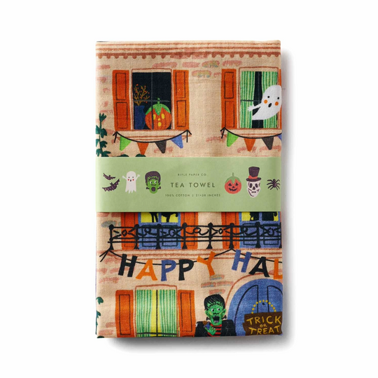 A folded cotton tea towel with a Halloween-themed illustration, wrapped in a light green paper band that reads 'TEA TOWEL' with small spooky icons. The towel features a beige brick building facade with orange window shutters, a ghost, a pumpkin, a frog, and 'HAPPY HALLOWEEN' bunting.