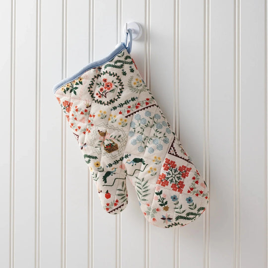 Decorative oven mitt with floral and nature-themed patterns hanging on a white wooden panel wall.