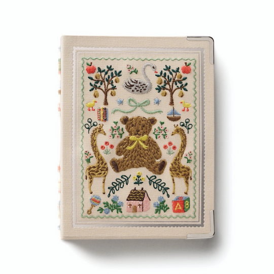 A decorative hardcover photo album book with a cream-colored cover featuring a colorful embroidery of a teddy bear, apples, birds, and small motifs. 
