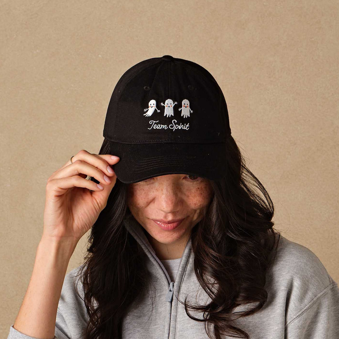 A woman wearing a black baseball cap with white embroidered cartoon ghosts and the text 'Team Spirit', paired with a light gray half-zip sweatshirt, posing against a textured beige background.
