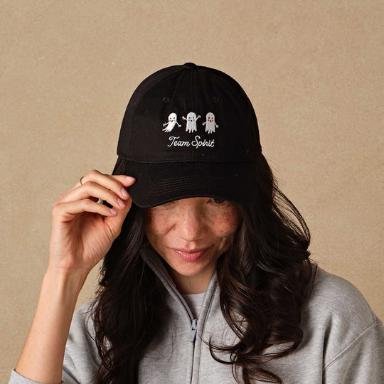 A woman wearing a black baseball cap with white embroidered cartoon ghosts and the text 'Team Spirit', paired with a light gray half-zip sweatshirt, posing against a textured beige background.