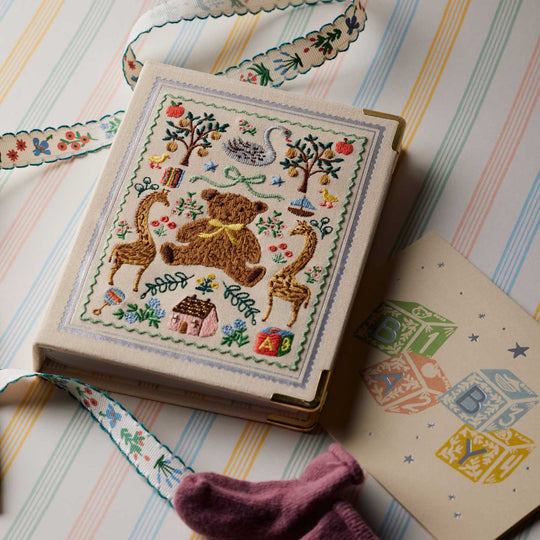 A decorative hardcover photo album book with a cream-colored cover featuring a colorful embroidery of a teddy bear, apples, birds, and small motifs. 